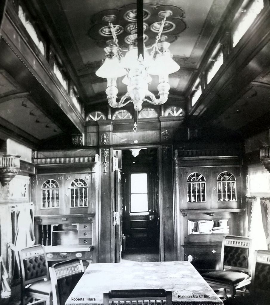 Historic photograph of the Interior view of Austin Corbin's private pullman railroad car featuring ornate wood paneling, a chandelier, and a dining area with a table and cushioned chairs.